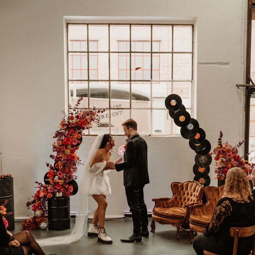 Bride in off shoulder tulle short wedding dress with church length veil and white platform Dr Martens wedding shoes laughing with groom in wet look black grooms suit with leopard print shirt and pink and orange boutonniere standing by vinyl wedding arch 