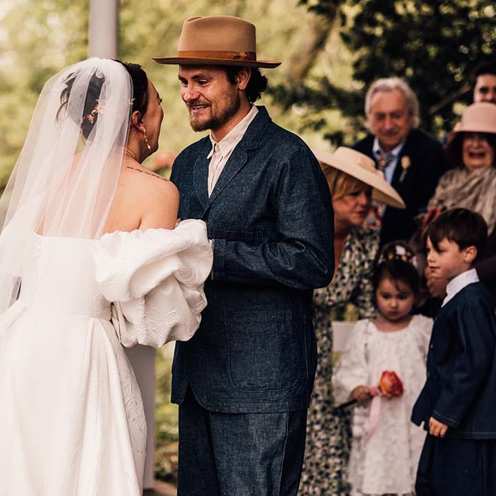 Bride in statement wedding dress leaning in for a kiss with the groom wearing stylish hat
