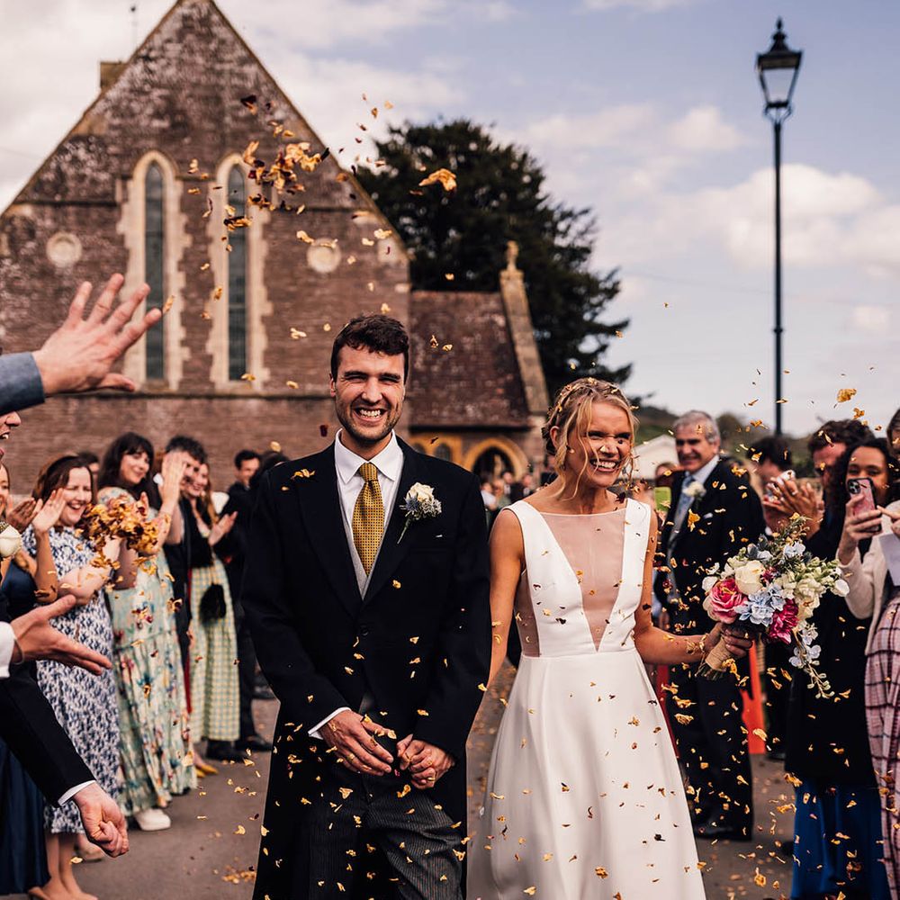 Groom in morning suit with yellow tie walking hand in hand with the bride to colourful confetti exit 