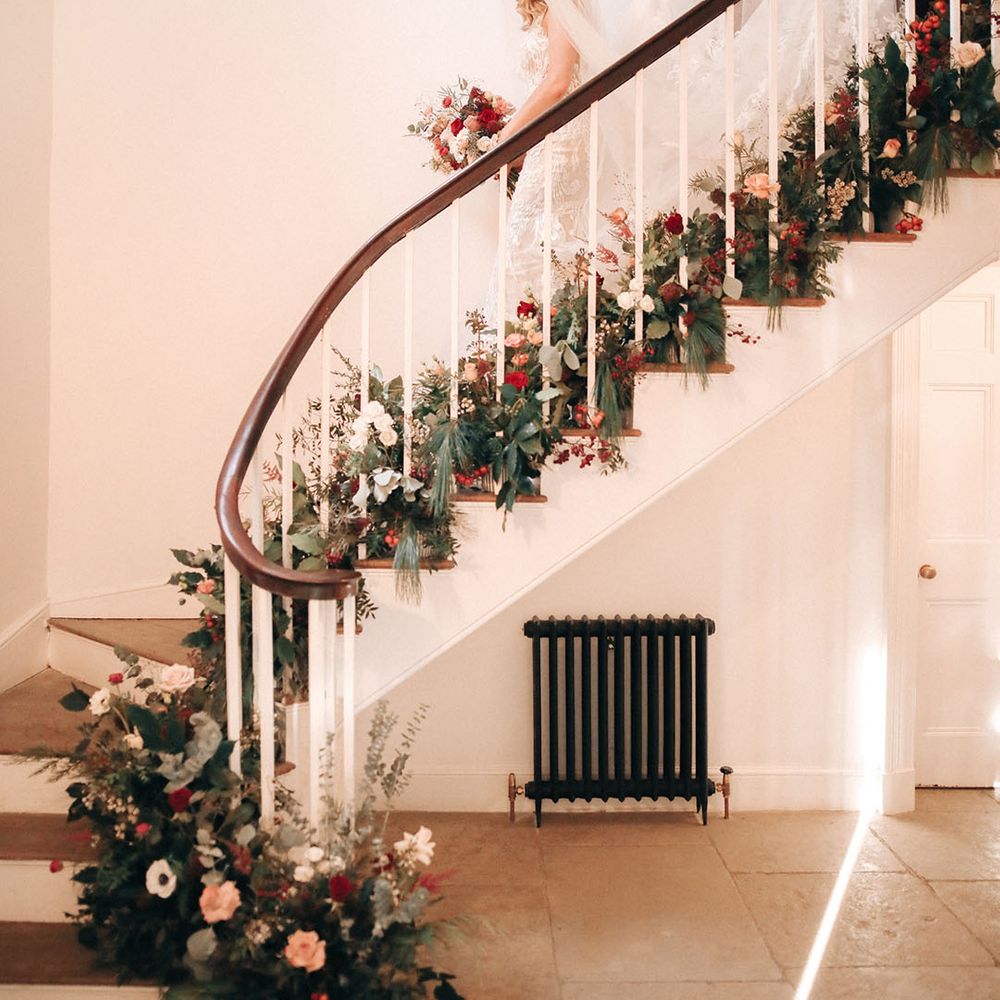 The bride and bridesmaids walk down the stairs together covered in festive seasonal winter flowers 