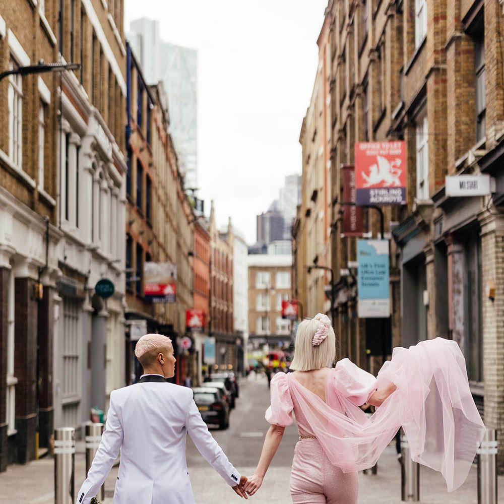 Two brides walking through Shoreditch holding hands in a pink jumpsuit with Watteau train and white tuxedo jacket 