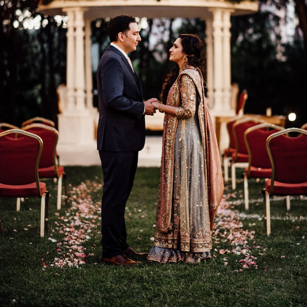 Bride & groom stand between chairs of petal line aisle as floral covered bandstand can be seen in the background