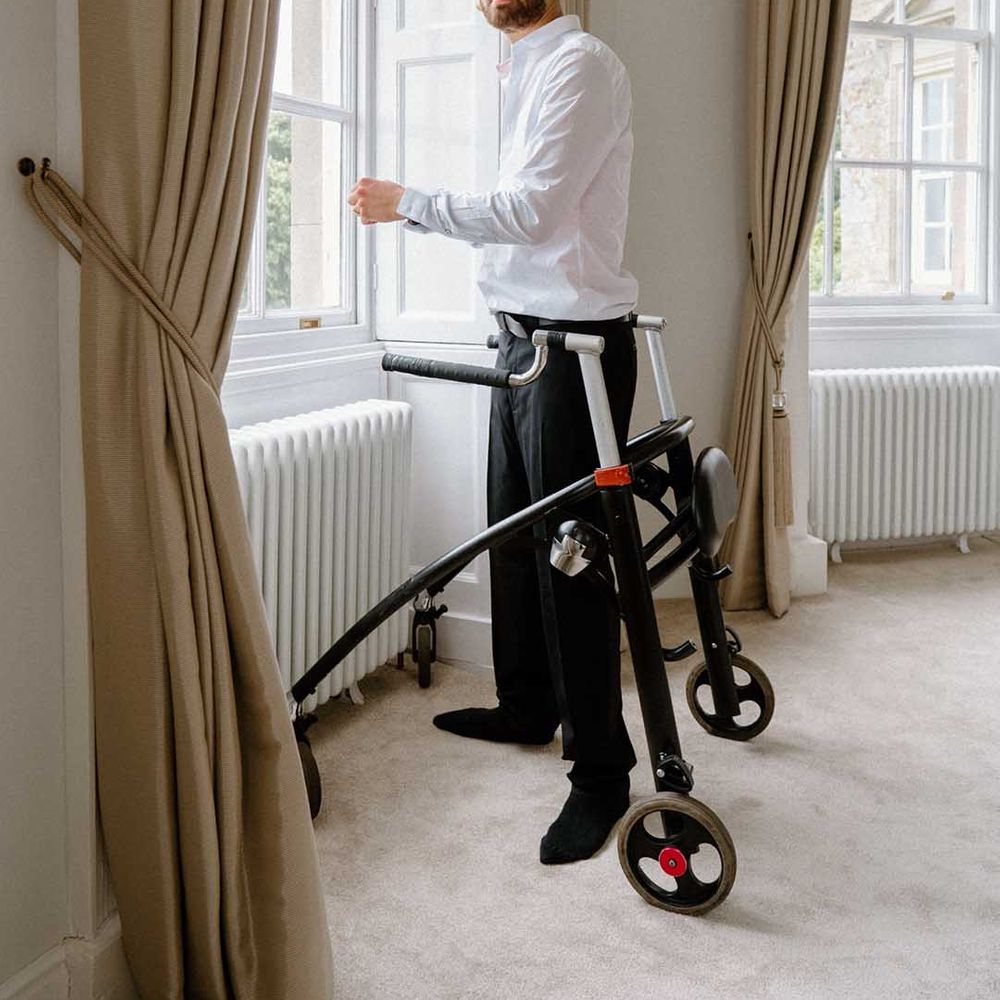 Disabled groom in a tuxedo standing at the window of the country house wedding venue with his walking frame 