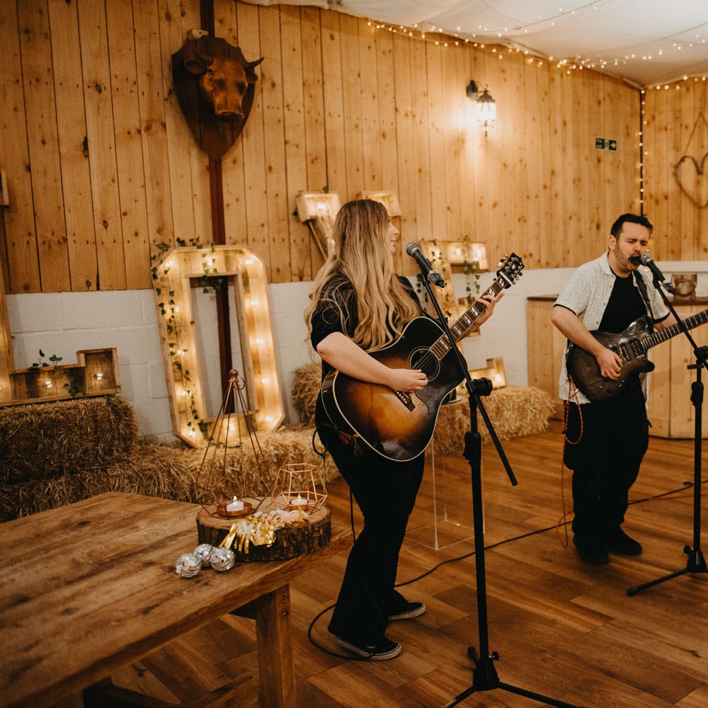 Two guitarists sing at microphones during rustic wedding ceremony in Lancashire with fairy lights, wooden tables and hay bales 