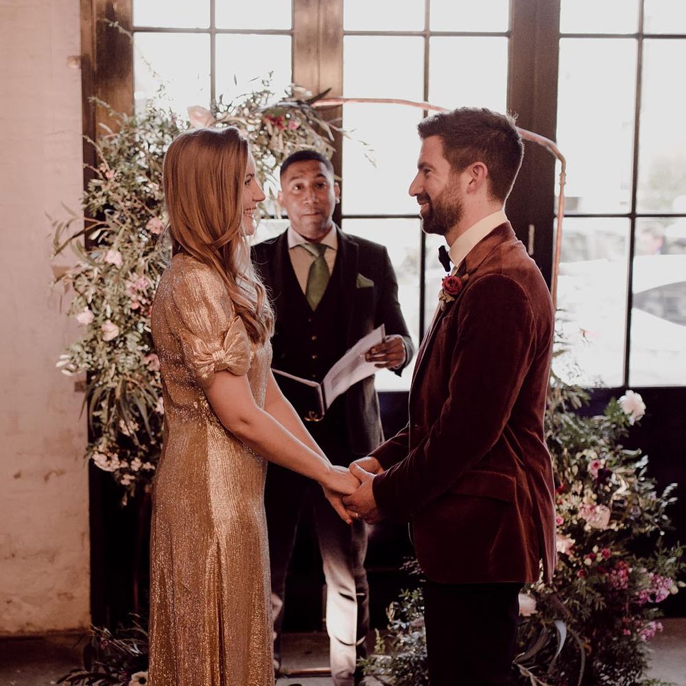 Groom in burgundy velvet suit jacket holds hands with bride in sparkly gold wedding dress with bow sleeves at the altar in front of frame with flower decor 
