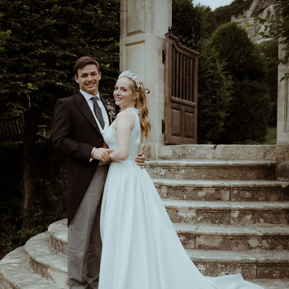 Bride smiles in pleated princess wedding dress with groom in morning suit after handfasting ceremony at Owlpen Manor 