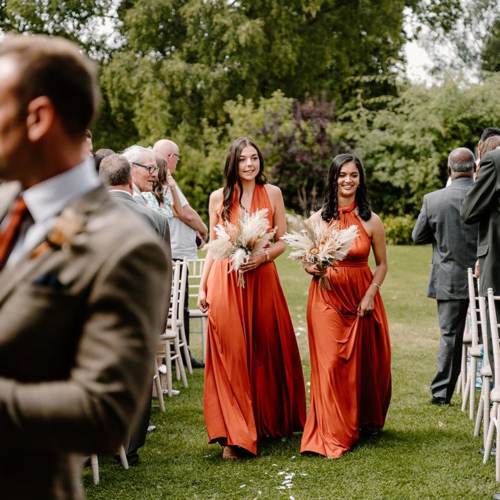 Bridesmaids in orange dresses walk down the aisle for the outdoor ceremony 