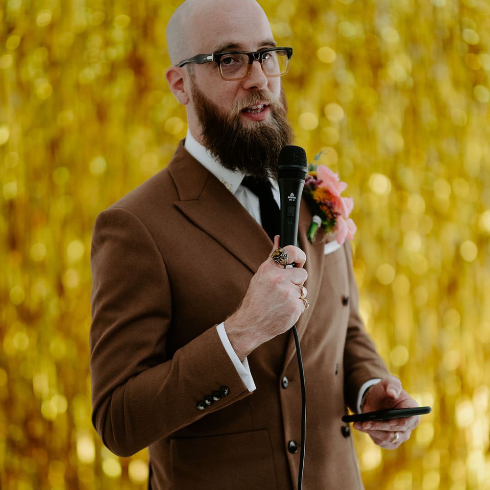 Groom wears brown retro suit and stands in front of gold streamers wedding decor 