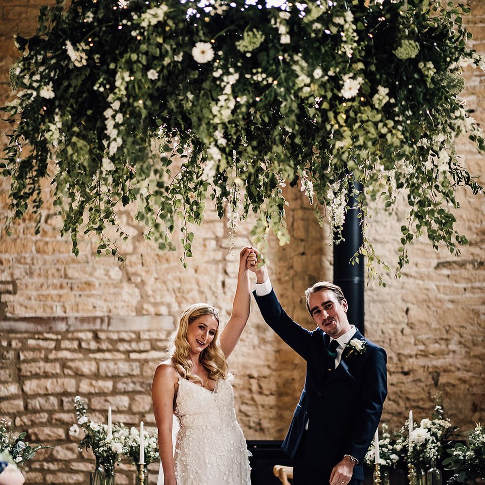 Foliage and white flower cloud hanging in the barn venue as the bride and groom raise their arms in celebration of their marriage 