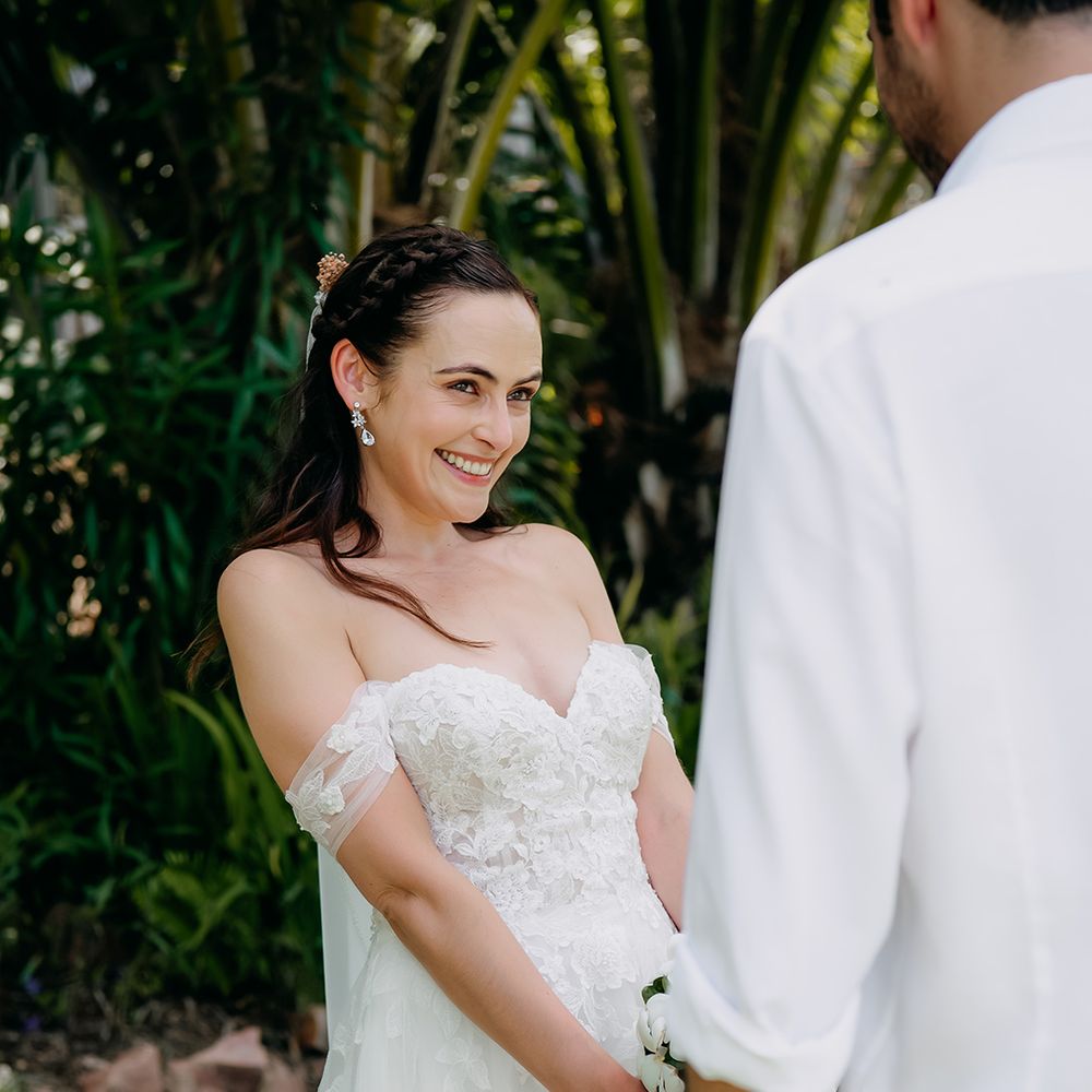 Bride in off the shoulder lace wedding dress with sweetheart neckline and lace floral details holding white tropical bouquet 