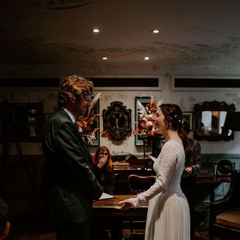 Groom in dark suit with orange tie and autumnal boutonniere and bride in long sleeve lace vintage wedding dress and closed toe satin heels holding hands at the alter 
