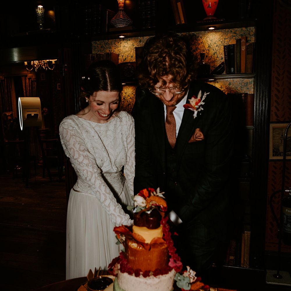 Bride in long sleeve lace vintage wedding dress with pearl earrings and pearl hair accessories and groom in dark suit with orange tie, orange pocket square and autumnal boutonniere cutting their cheese board cake tower  