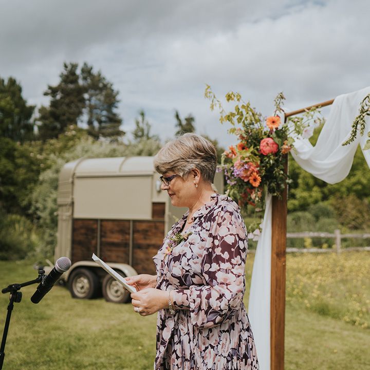 Wedding guest speaks during outdoor wedding ceremony in front of wooden arch finished with white drapes and colourful foliage 