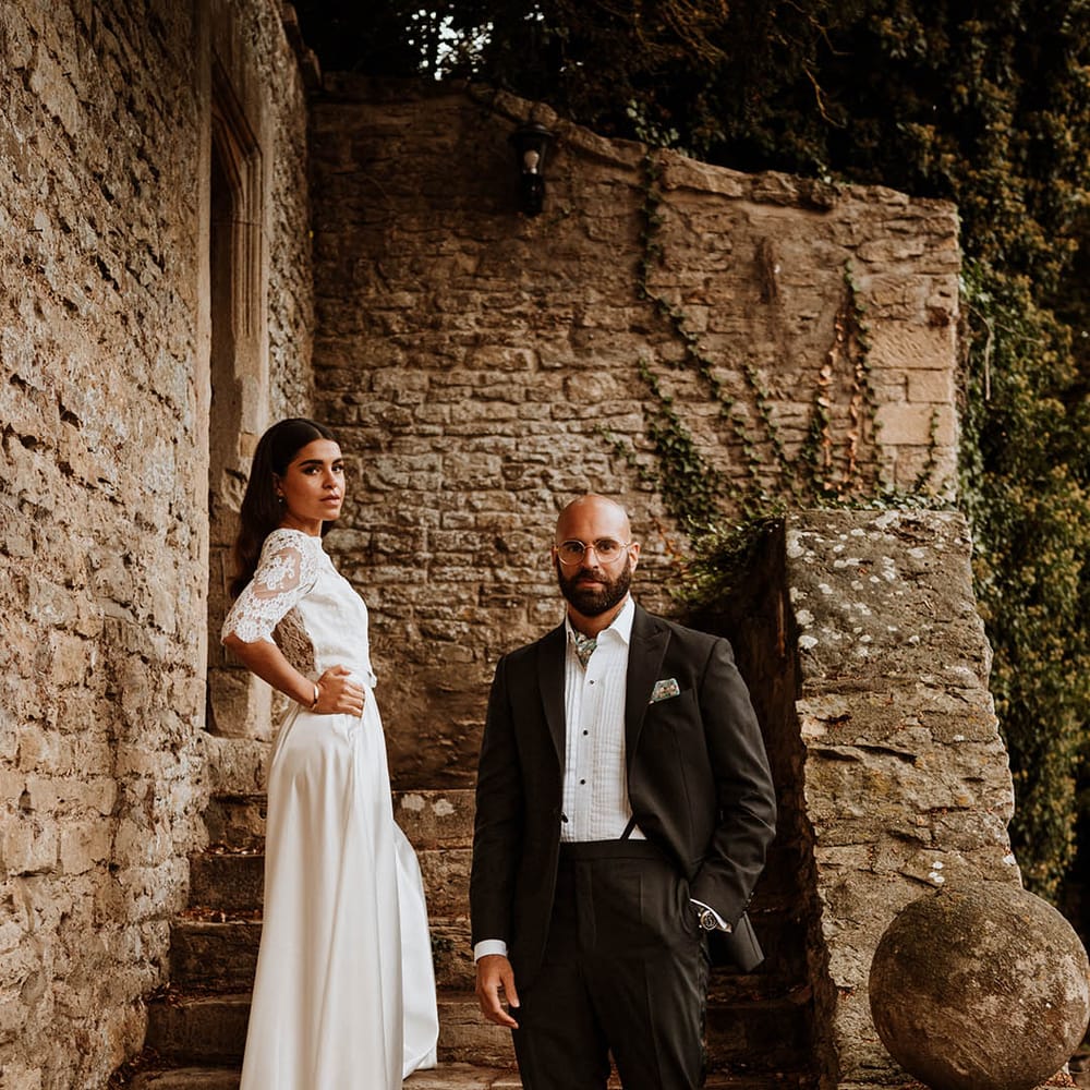 Bride in three quarter length sleeve wedding dress with lace detailing and full satin skirt posing with groom in classic black tuxedo with hand painted silk cravat and handkerchief on the stairs of glasshouse wedding venue