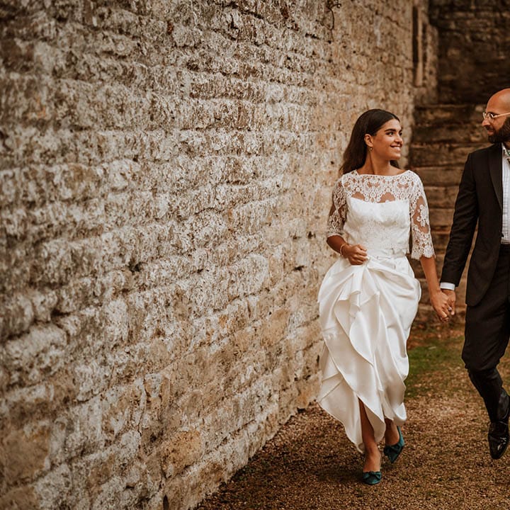 Groom in classic black tuxedo with hand painted silk cravat and handkerchief and bride in three quarter length sleeve wedding dress with lace detailing and full satin skirt and bottle green satin wedding shoes holding hands and walking around wedding venue