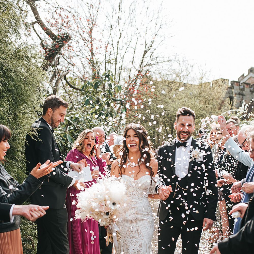 Bride in strapless boho fitted lace wedding dress with detachable sleeves having white confetti men with the groom in black tie 