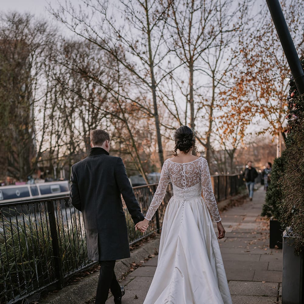 Bride in affeta material princess wedding dress with long lace sleeves  and cropped faux fur bridal cover up holding hands with groom in stylish black tuxedo with black bowtie walking around London