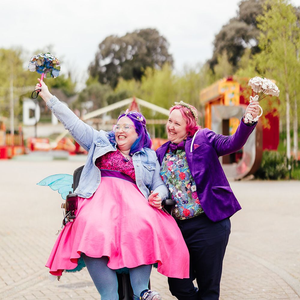 Bride in purple velvet blazer and black suit trousers with purple bowtie, pink patterned pocket square and colourful patterned shirt holding paper flower bouquet and bride in wheelchair wearing personalised denim jacket, pink wedding dress and blue tights holding colourful paper flower bouquet 