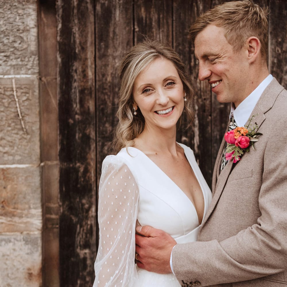 The bride in a plunging neckline wedding dress with long sleeves and pearl earrings smiles with the groom who wears a neutral coloured wedding suit with a floral tie from Peggy and Finn