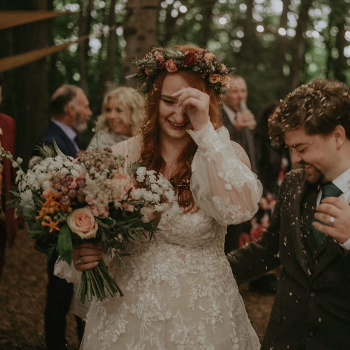 Bride wearing off the shoulder long sleeve boho wedding dress with flower crown walking with the groom as confetti is thrown over the guests 