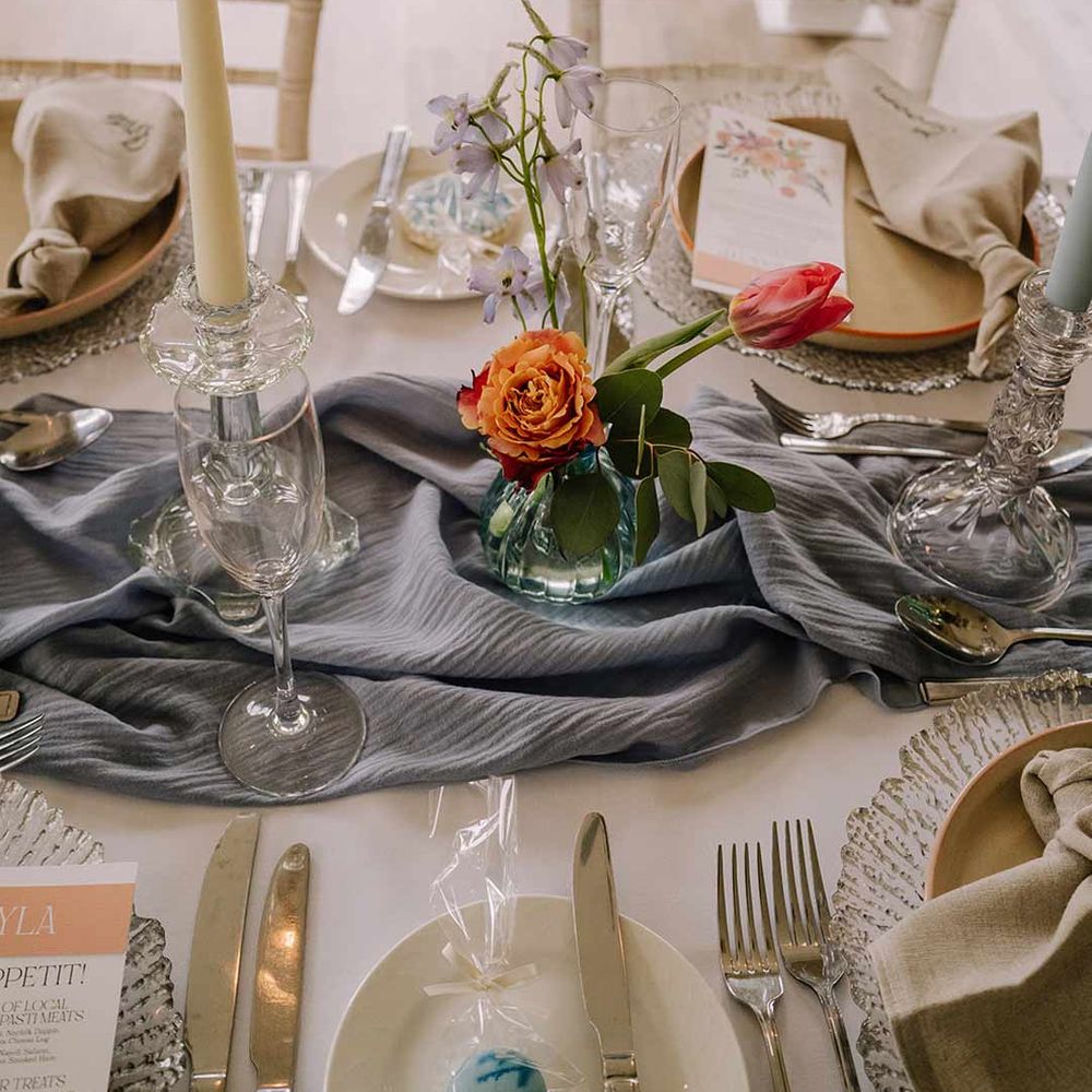 Cream tapered candle in glass candlestick holder on cornflower blue wedding table runner with bright blue glass bottles with dried flowers