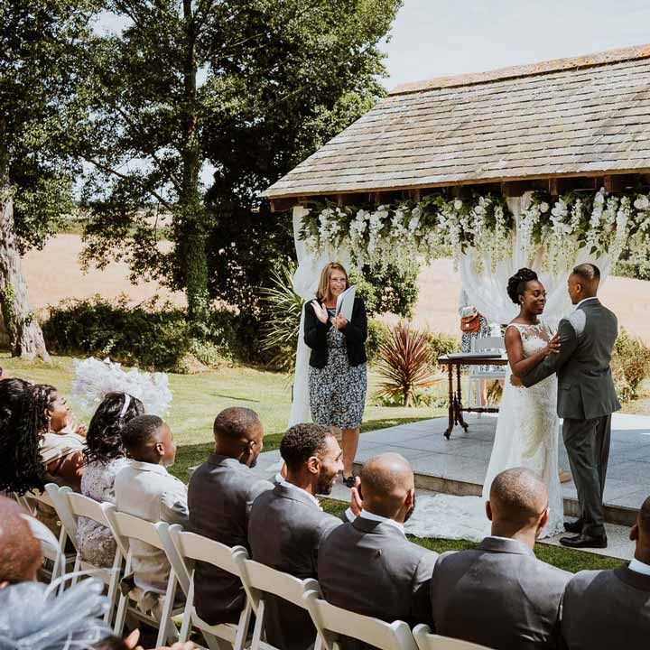 Bride in lace and mesh boatneck wedding dress with silver bridal hair accessories standing with groom in charcoal grey grooms suit, light grey patterned waistcoat, light grey patterned tie and large white rose and foliage boutonniere standing at the end of the alter of Tredudwell Manor with registrar 