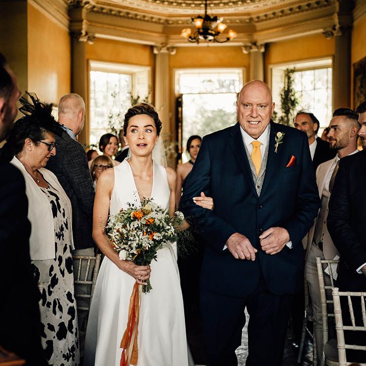 Father of the bride in a dark blue suit with yellow tie walks the bride down the aisle to meet the groom 