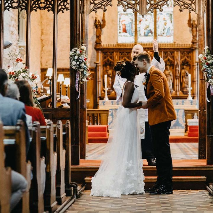 Bride in v neck sleeveless wedding dress with 3D floral applique train kissing groom in crushed burnt orange velvet grooms blazer, black bowtie and black suit trousers at the alter of Brinsop Court wedding venue
