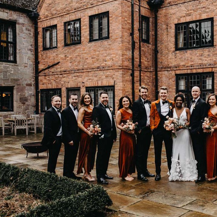 Wedding party wearing classic black groomsmen suits, one shoulder burnt orange bridesmaid dresses, crushed burnt orange velvet grooms blazer, black bowtie and black suit trousers and a v neck sleeveless wedding dress with 3D floral applique train all standing outside of Brinsop Court wedding venue