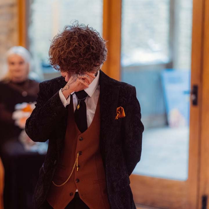 Bride in dark velvet bridal jacket with rast waistcoat and pocket square, black suit trousers `nd gold pocket watch wiping away a tear at the alter of Kingscote Barn Tetbury