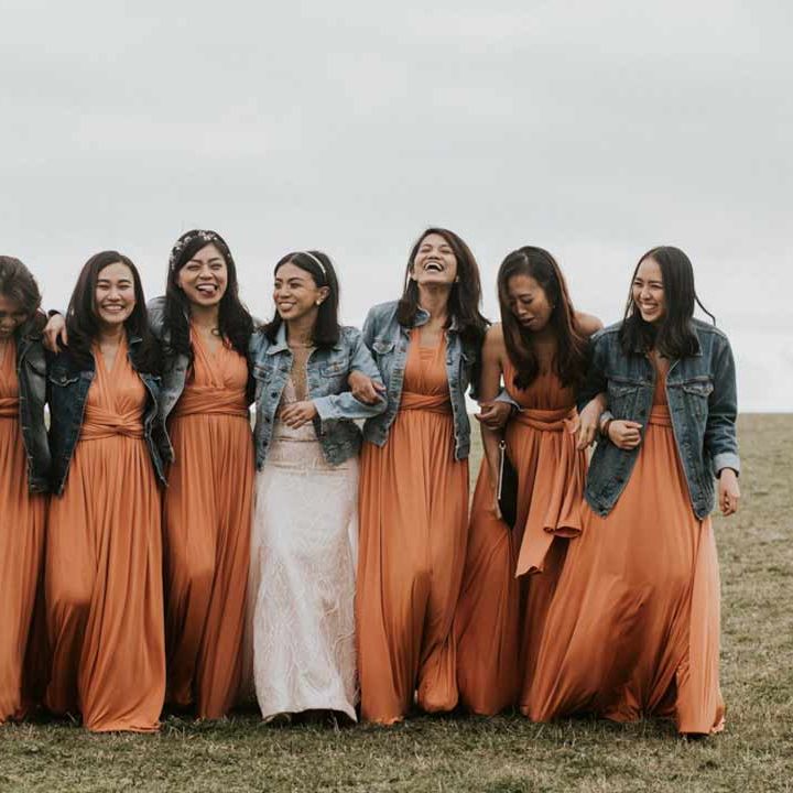 Bridesmaids in front wrap orange bridesmaid dresses and dark wash denim jackets walking with bride in lace wedding dress with denim bridal coverup