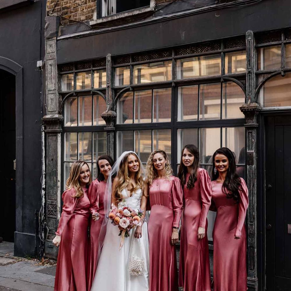 Bride in corset top satin wedding dress standing with bridesmaids in long sleeve satin pink bridesmaid dresses 