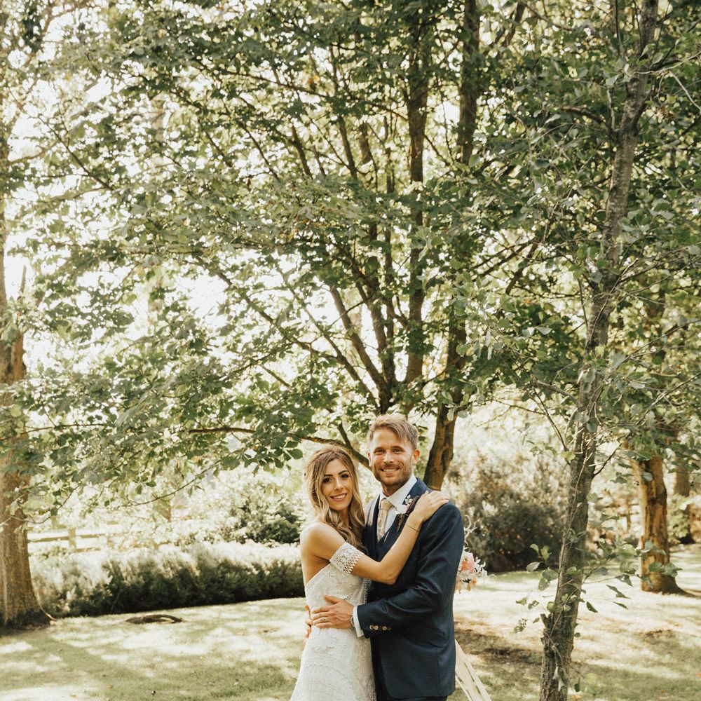 Boho bride in Clara Rue De Seine Dress hugs Groom in navy bespoke suit in garden at Tythe Barn Wedding