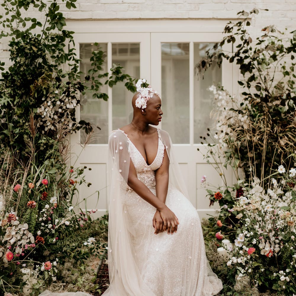 Black bride with alopecia sitting at the altar in a blush pink Julita Bridal London wedding dress with cape and Ophelia Ren Bridal headdress 