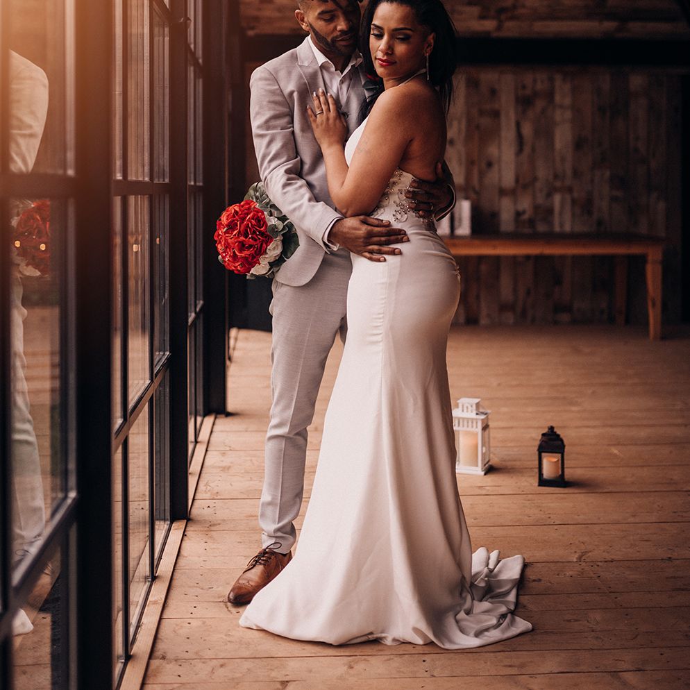 Groom wraps arms around bride as he looks at her, and bride holds bouquet of roses 