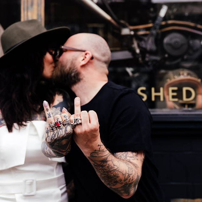 Tattooed bride and groom holding their wedding fingers up whilst kissing 