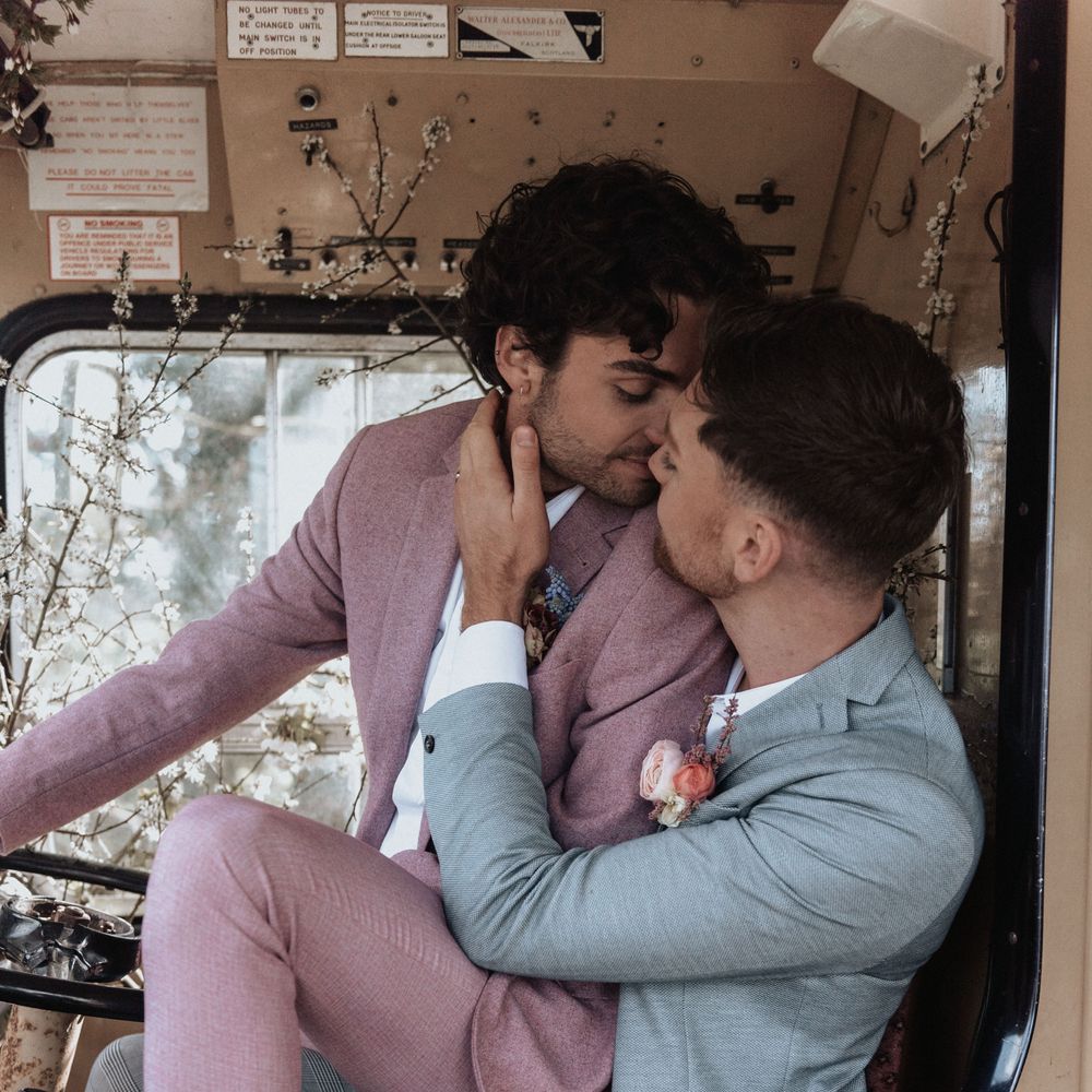Groom sits on groom whilst wearing pastel suits and kissing on wedding day
