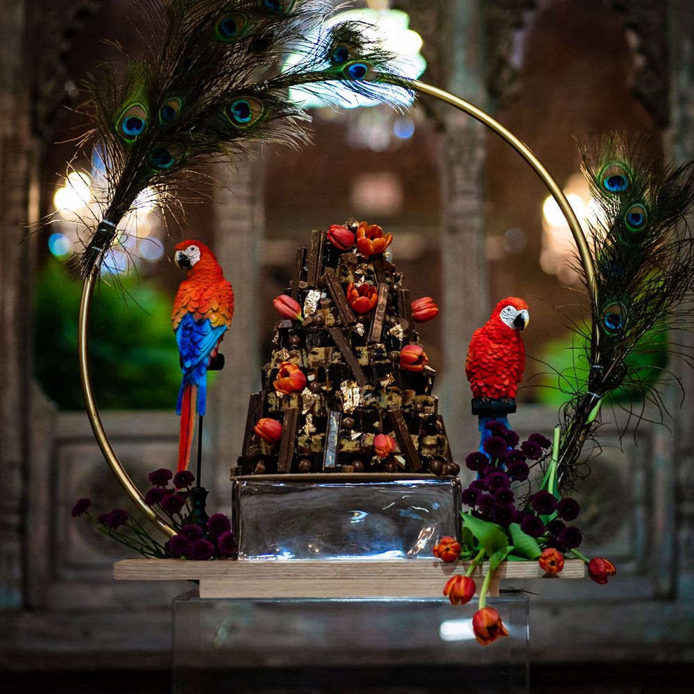 Chocolate wedding cake with chocolate biscuit fingers and red tulip decor on a hoop cake stand decorated with peacock feathers and parrots 
