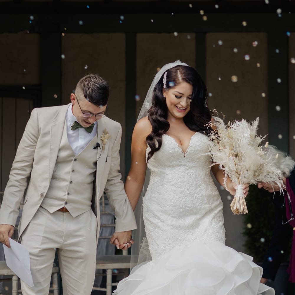 Bride in a lace fitted fishtail wedding dress with long wavy hair holding hands with her groom in a three piece beige suit 