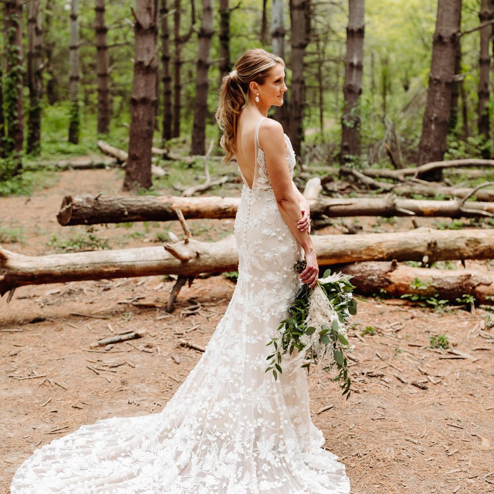 Bride in white low v back embroidered Made With Love wedding dress stands in woodland holding white, pink and green bridal bouquet at backyard wedding