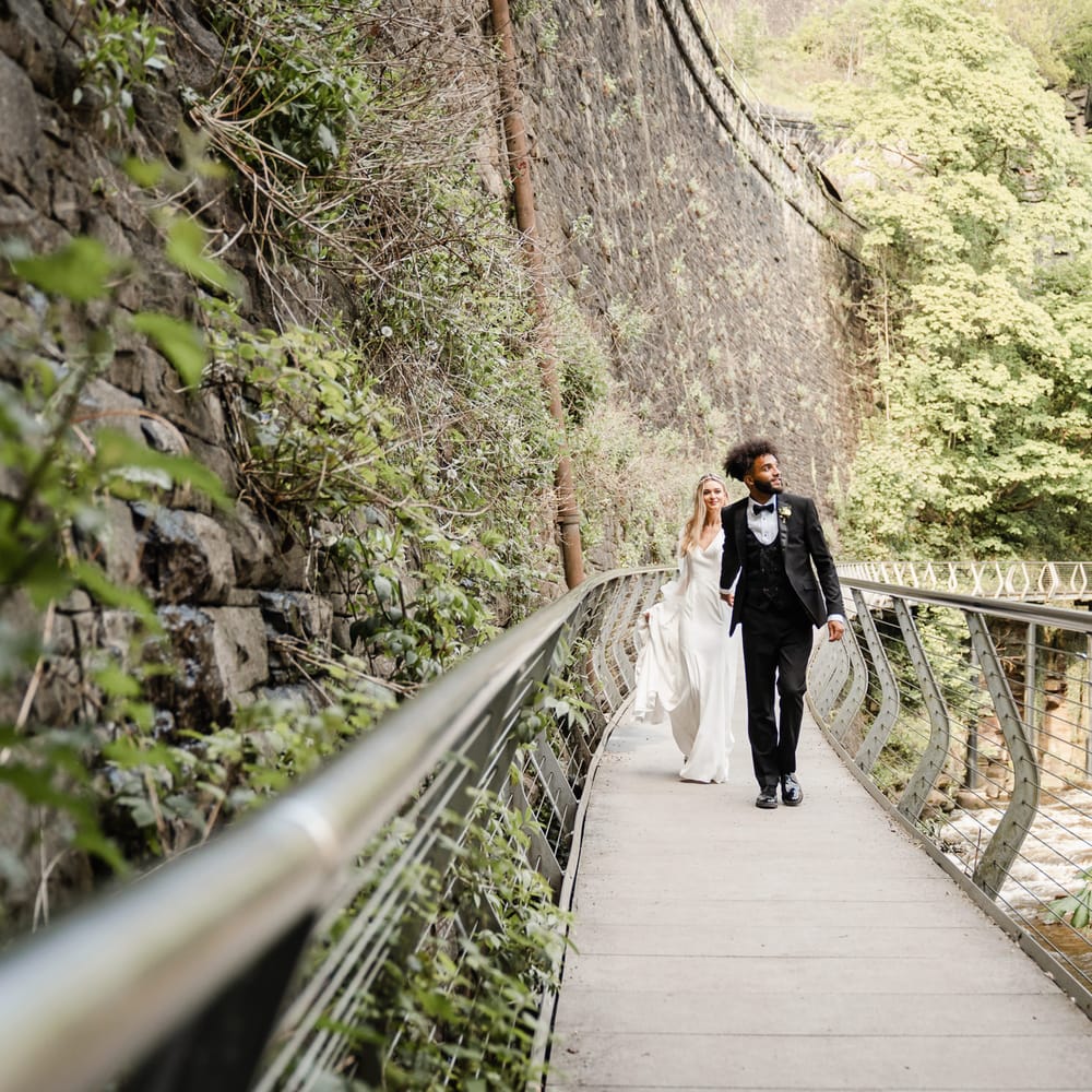 Portrait of a groom in a black tuxedo with horseshoe waistcoat holding hands with this bride on a bridge 