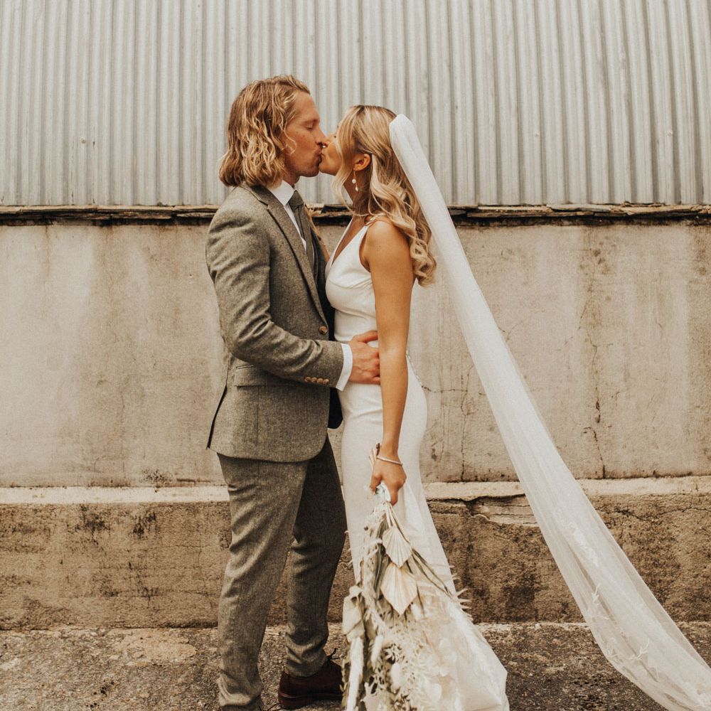Bride in white Made With Love wedding dress and embroidered veil holding white and green dried bridal bouquet kisses groom in sage green suit and brown shoes outside after wedding ceremony at Anran Devon