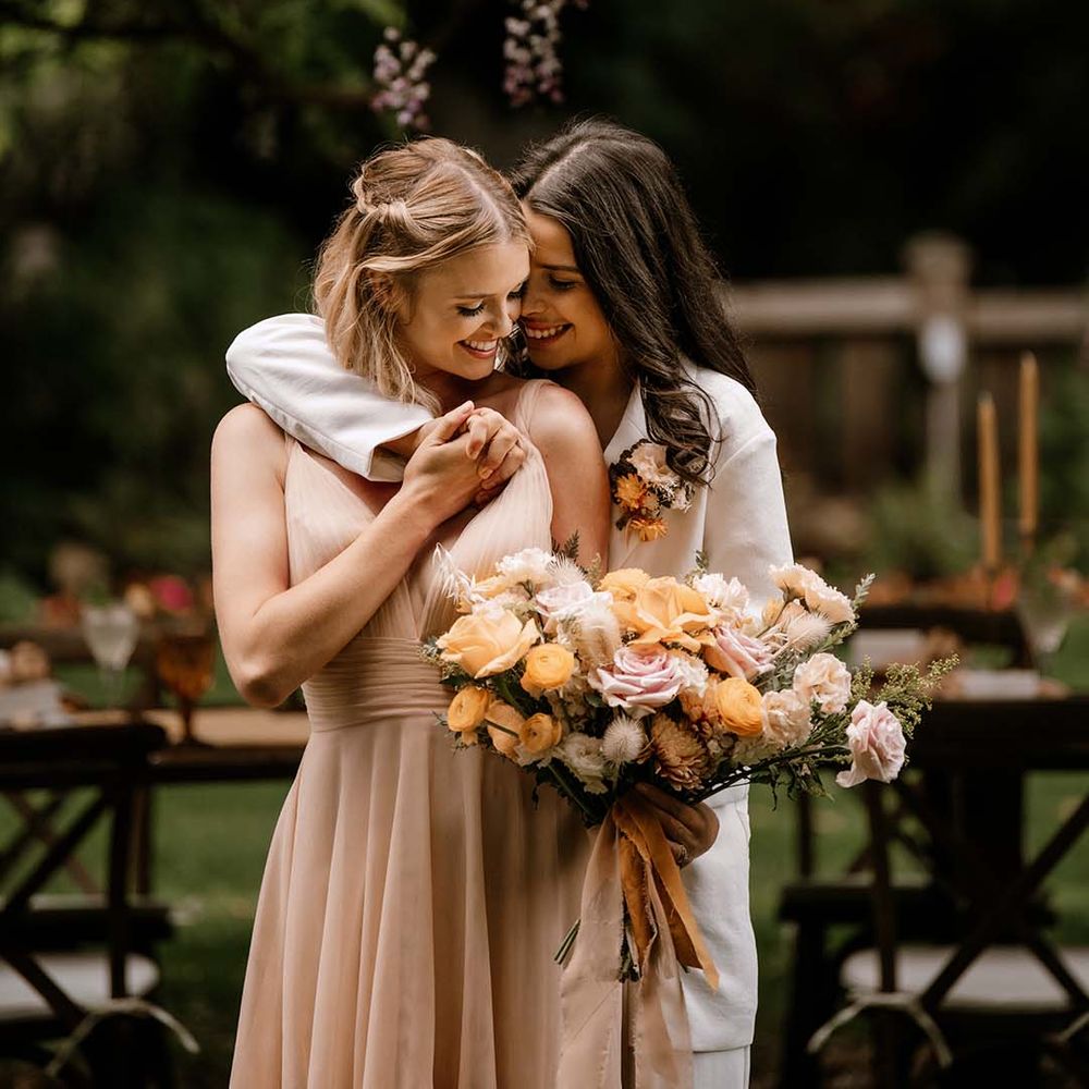 Bride in a white suit embracing her bride in a blush pink wedding dress as she holds a peeled back rose wedding bouquet 