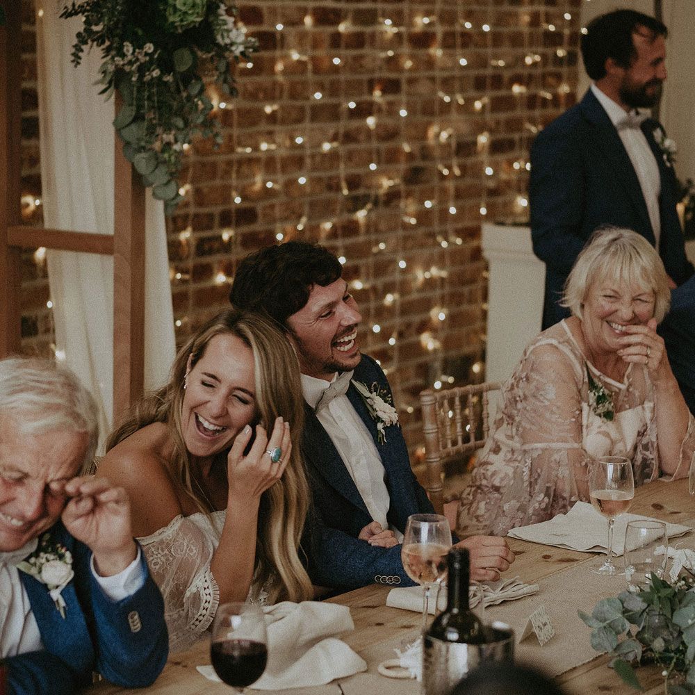 Bride in Grace Loves Lace bardot wedding dress and groom in blue jacket and bow tie laugh with wedding guests as they sit at top table in front of string fairy lights at Isle of Wight wedding with macrame wedding decor