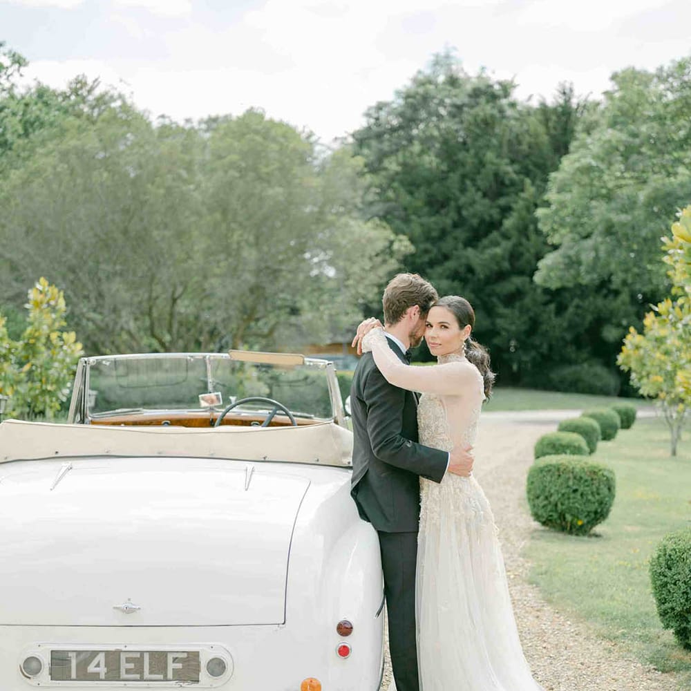 Bride in a romantic lace wedding dress with long sleeves embracing her husband in a tuxedo by their vintage wedding car 