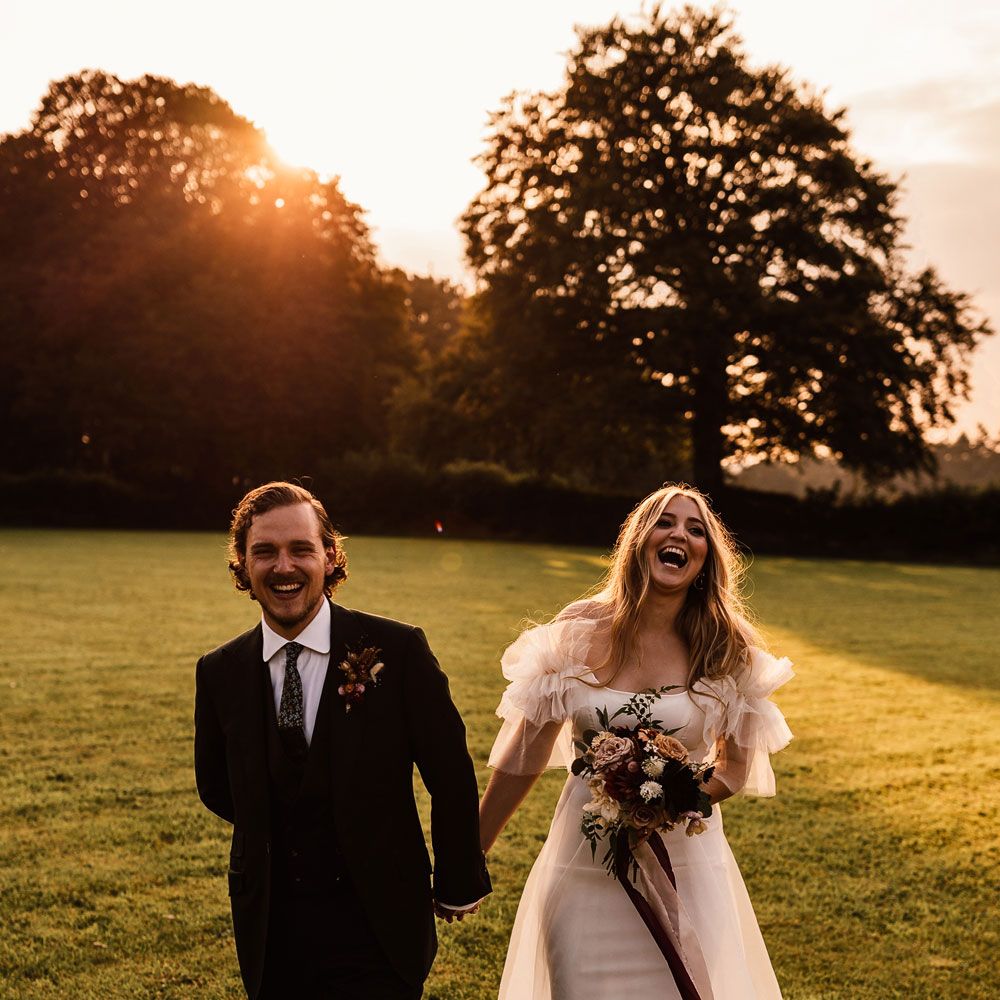Bride in whit Halfpenny London Mayfair dress with ruffle sleeves holding mixed bridal bouquet with burgundy ribbons walks through garden holding hands with groom in three piece suit and buttonhole during holden hour at wedding in Cornwall