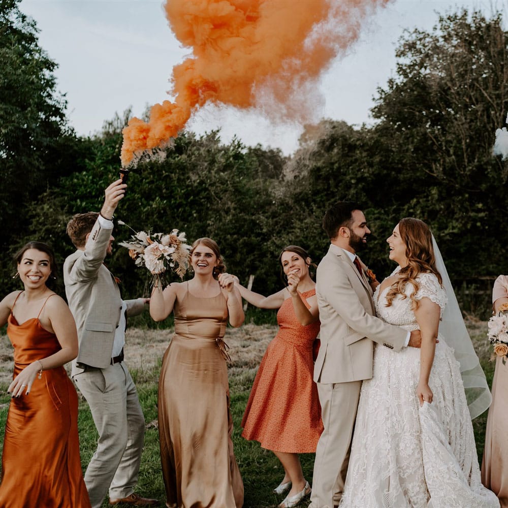 Bride in homemade lace wedding dress holds hands with groom in beige three piece suit as they stand with groomsmen and bridesmaids in field whilst holding orange and white smoke bombs