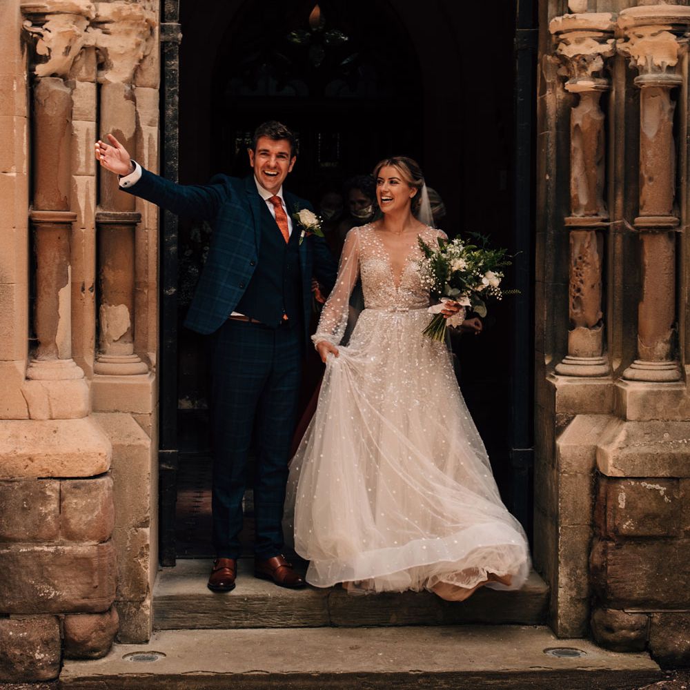 Groom in blue checked suit throws out his arms as he leaves church with bride in mesh long sleeve Julie Vino wedding dress and veil after wedding ceremony