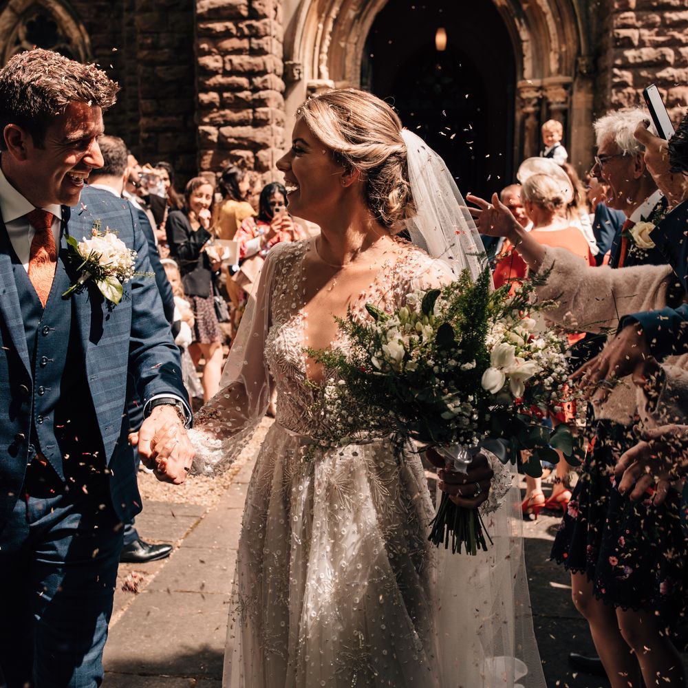 Groom in blue three piece checked suit smiles as he holds hands with bride in long sleeve embroidered Julie Vino wedding dress and veil whilst guests throw confetti
