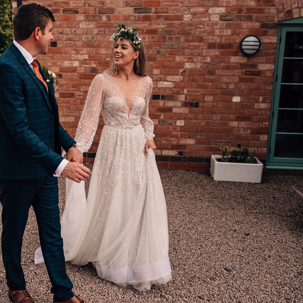 Bride in long mesh sleeve Julie Vino wedding dress and flower crown walks across drive with groom in blue checked three piece suit and floral button hole